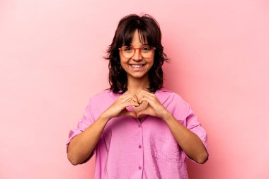 Pembe arka planda izole edilmiş genç İspanyol kadın gülümsüyor ve elleriyle kalp şeklini gösteriyor..