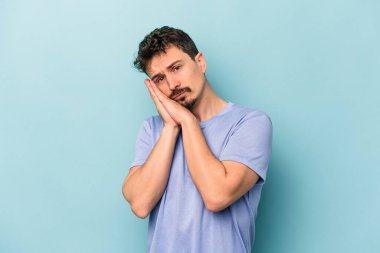 Mavi arka planda izole edilmiş genç beyaz adam esniyor. Ağzı eliyle kapatan yorgun bir hareket sergiliyor..