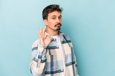 Kafkasyalı genç bir adam mavi arka planda izole edilmiş bir şekilde göz açıp kapayana kadar iyi bir el hareketi yapıyor..