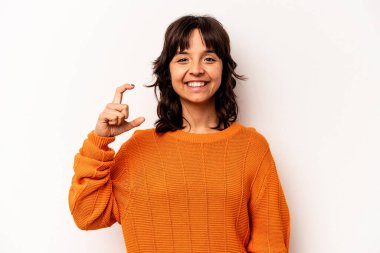 Beyaz arka planda izole edilmiş genç İspanyol kadın işaret parmaklarıyla küçük bir şey tutuyor, gülümsüyor ve kendinden emin..