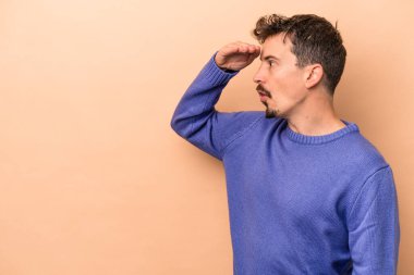 Beyazlı genç bir adam bej arka planda izole edilmiş, uzaklara bakıyor ve elini alnına koyuyor..