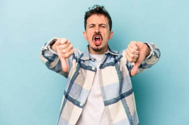 Mavi arka planda izole edilmiş genç beyaz bir adam baş parmağını aşağı gösteriyor ve hoşlanmadığını ifade ediyor..