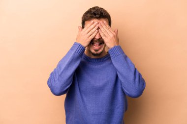 Beyaz tenli, bej arka planda izole edilmiş genç bir adam elleriyle gözlerini kapatıyor, gülümseyerek büyük bir sürpriz bekliyor..