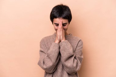 Beyaz tenli genç bir kadın, bej arka planda izole edilmiş, el ele tutuşmuş, ağız kenarında dua ediyor, kendinden emin..