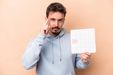 Kafkasyalı genç bir adam bej arka planda izole edilmiş bir yapboz tutuyor. Parmağıyla tapınağı işaret ediyor, düşünüyor, bir göreve odaklanıyor..