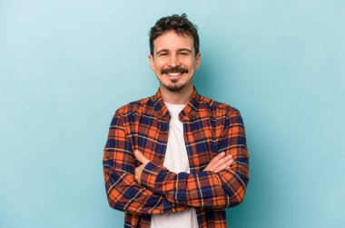 Mavi arka planda kendini güvende hisseden, kararlılıkla kollarını kavuşturan genç beyaz bir adam..