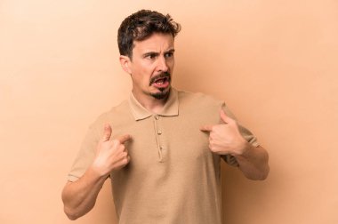 Beyaz tenli genç adam bej arka planda izole edildi. Parmakla göstererek şaşırdı, gülümsedi..