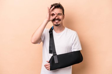 Kırık kollu genç beyaz adam bej arka planda izole edilmiş. Heyecanlı. Göze göz hareketi yapıyor..