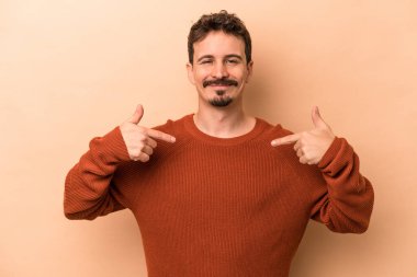 Beyazlı genç bir adam bej arka planda izole edilmiş. Elle bir gömlek fotokopi alanını işaret ediyor. Gururlu ve kendinden emin.