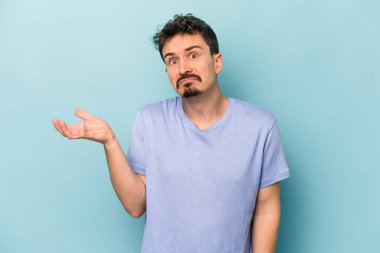 Kafkasyalı genç adam mavi arka planda izole edilmiş sorgulama jestinde şüphe ediyor ve omuz silkiyor..