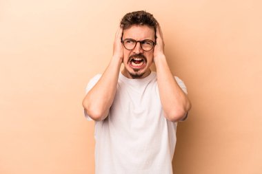Beyazlı genç bir adam bej arka planda izole edilmiş kulakları kulaklarıyla kapatıyor çok fazla ses duymamaya çalışıyor..