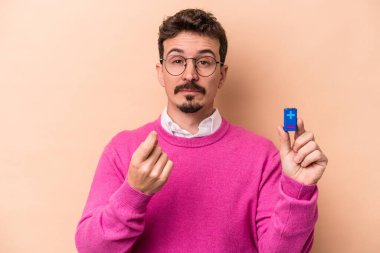 Elinde bataryaları olan genç beyaz bir adam bej arka planda izole edilmiş ve seni parmağıyla işaret ediyor sanki davet ediyormuş gibi..