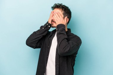 Mavi arka planda izole edilmiş genç beyaz adam elleriyle gözlerini kapatmaktan korkuyor..