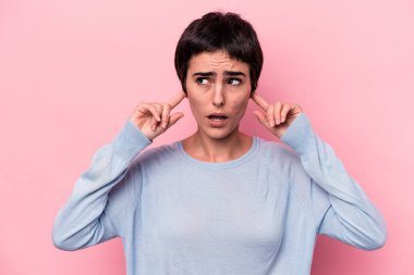 Pembe arka planda izole edilmiş genç beyaz bir kadın parmaklarıyla kulaklarını kapatıyor, gürültülü bir ortam yüzünden stresli ve çaresiz..