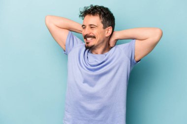 Kafkasyalı genç adam mavi arka planda izole edilmiş. Kendine güveniyor, elleri başının arkasında..