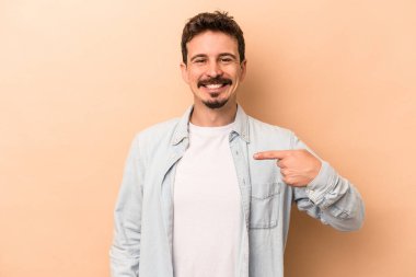 Beyazlı genç bir adam bej arka planda izole edilmiş. Elle bir gömlek fotokopi alanını işaret ediyor. Gururlu ve kendinden emin.