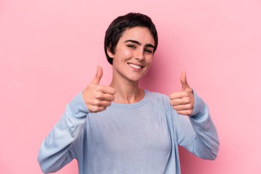 Pembe arka planda izole edilmiş genç beyaz kadın iki parmağını da kaldırıyor, gülümsüyor ve kendinden emin..