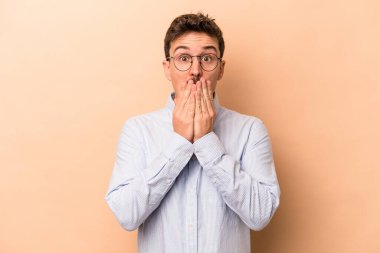 Beyaz tenli genç adam bej arka planda izole edilmiş şok olmuş, ağzı ellerle kaplanmış, yeni bir şey keşfetmeye hevesli..