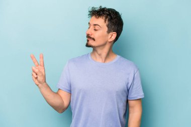 Kafkasyalı genç adam mavi arka planda izole edilmiş neşeli ve kaygısız parmaklarıyla barış sembolü gösteriyor..