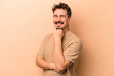 Beyaz tenli genç adam bej arka planda izole edilmiş mutlu ve kendinden emin gülümsüyor, elleriyle çenesine dokunuyor..