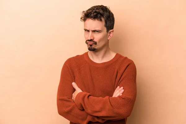Young caucasian man isolated on beige background frowning face in displeasure, keeps arms folded.
