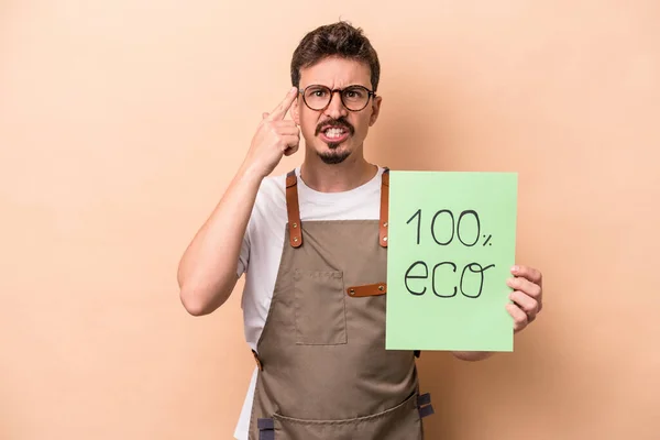 Young caucasian gardener man holding 100% eco placard isolated on beige background showing a disappointment gesture with forefinger.