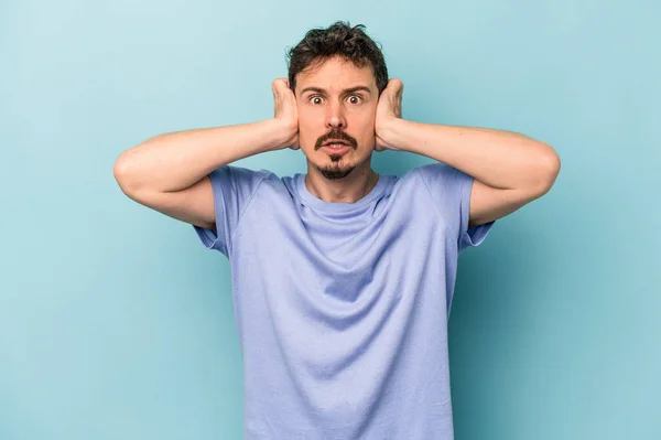 Young caucasian man isolated on blue background covering ears with hands trying not to hear too loud sound.