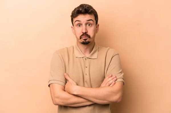 Young caucasian man isolated on beige background blows cheeks, has tired expression. Facial expression concept.