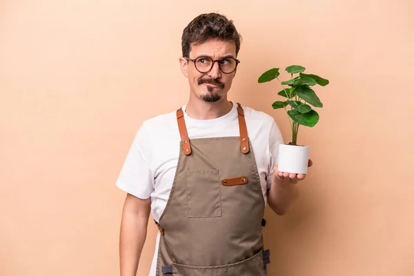 Young caucasian gardener man holding a plant isolated on beige background confused, feels doubtful and unsure.