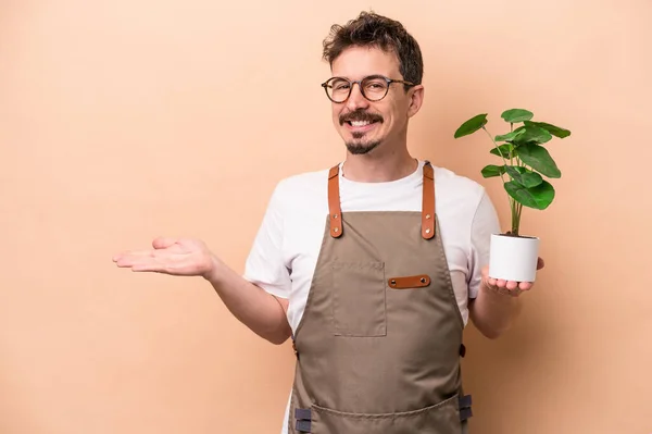 Young caucasian gardener man holding a plant isolated on beige background showing a copy space on a palm and holding another hand on waist.