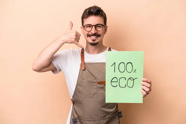Young caucasian gardener man holding 100% eco placard isolated on beige background showing a mobile phone call gesture with fingers.