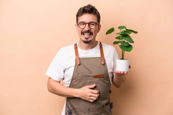 Young caucasian gardener man holding a plant isolated on beige background laughing and having fun.