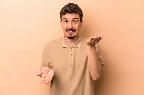 Young Caucasian Man Isolated Beige Background Makes Scale Arms Feels — Stock Photo, Image
