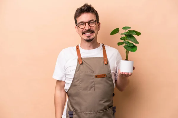 Young caucasian gardener man holding a plant isolated on beige background happy, smiling and cheerful.