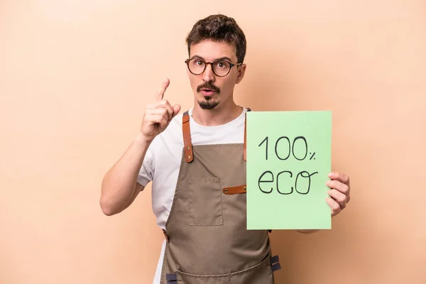 Young caucasian gardener man holding 100% eco placard isolated on beige background having an idea, inspiration concept.