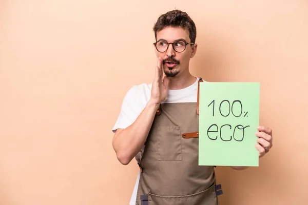 Young caucasian gardener man holding 100% eco placard isolated on beige background is saying a secret hot braking news and looking aside