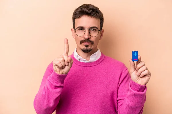 Mladý Běloch Drží Baterie Izolované Béžovém Pozadí Ukazující Číslo Jedna — Stock fotografie