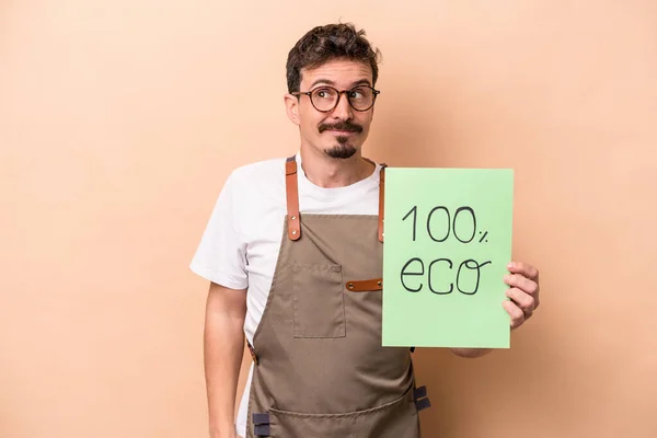 Young caucasian gardener man holding 100% eco placard isolated on beige background dreaming of achieving goals and purposes