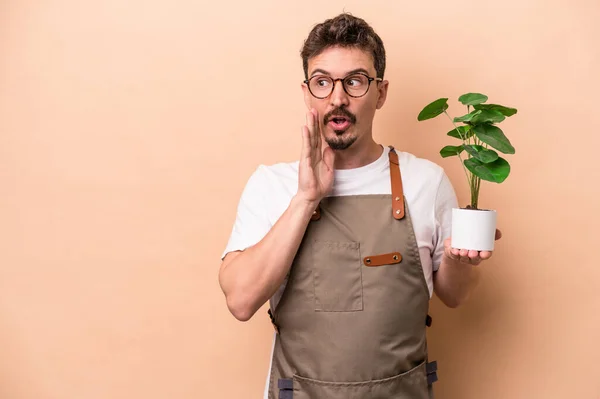 Young caucasian gardener man holding a plant isolated on beige background is saying a secret hot braking news and looking aside