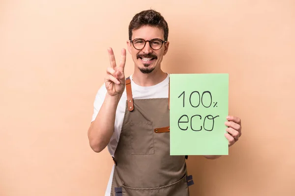 Young caucasian gardener man holding 100% eco placard isolated on beige background showing number two with fingers.