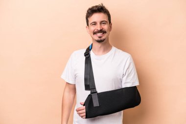 Kırık kollu genç beyaz adam bej arka planda izole edilmiş mutlu, güler yüzlü ve neşeli..