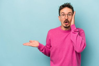 Mavi arka planda izole edilmiş genç beyaz adam, avuç içi fotokopi alanını elinde tutmaktan etkilenmiş..