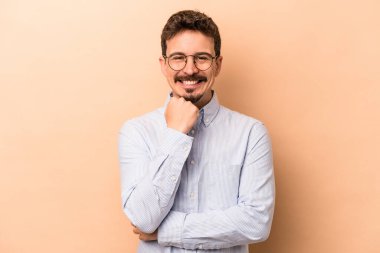Beyaz tenli genç adam bej arka planda izole edilmiş mutlu ve kendinden emin gülümsüyor, elleriyle çenesine dokunuyor..