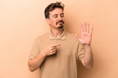 Beyaz tenli genç adam bej arka planda izole edilmiş. Gülümsüyor. Beş numarayı parmaklarıyla gösteriyor..