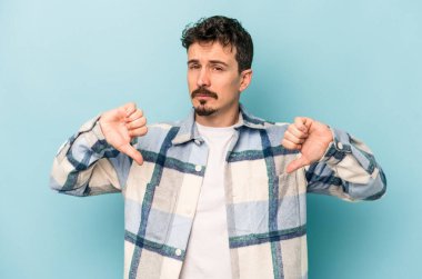 Mavi arka planda izole genç beyaz adam baş parmağını aşağı gösteriyor, hayal kırıklığı konsepti.