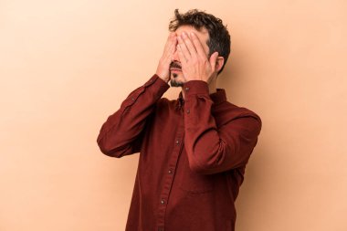 Beyazlı genç bir adam bej arka planda izole edilmiş, elleriyle gözlerini kapatmaktan korkuyor..