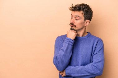 Beyazlı genç adam bej arka planda izole edilmiş şüpheli ve şüpheci bir ifadeyle yana bakıyor..