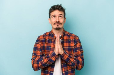 Mavi arka planda izole edilmiş genç beyaz bir adam el ele tutuşup, ağzının kenarında dua ediyor, kendinden emin..