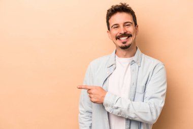 Beyaz tenli genç adam bej arka planda izole edilmiş gülümsüyor ve boş alanda bir şeyler gösteriyor..