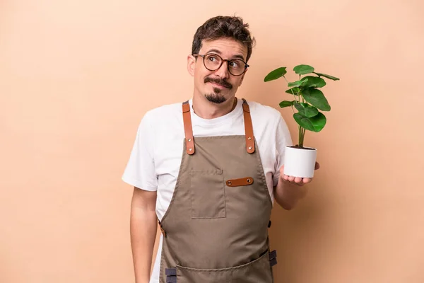 Young caucasian gardener man holding a plant isolated on beige background dreaming of achieving goals and purposes